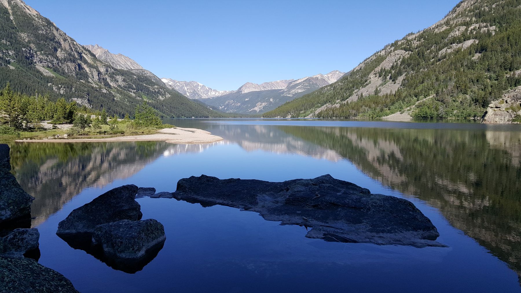 Boulders in the still shallows of Mystic Lake, mirroring forested slopes and snow-streaked peaks