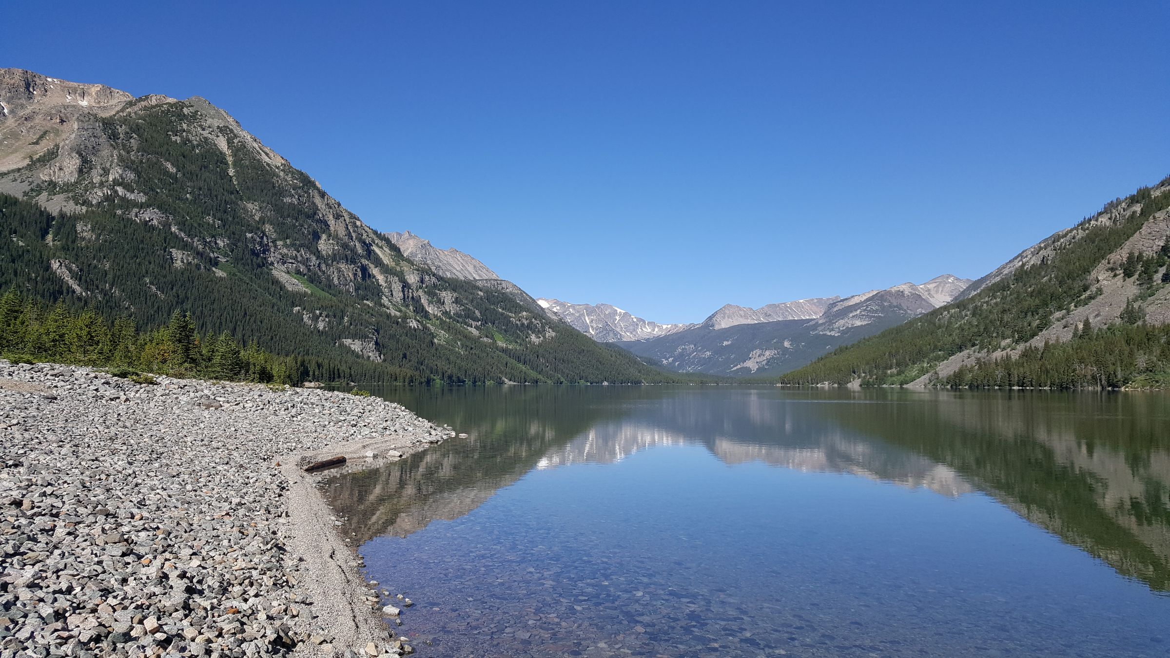 The gravel shore of Mystic Lake, with pine slopes and the Beartooth peaks mirrored in still water